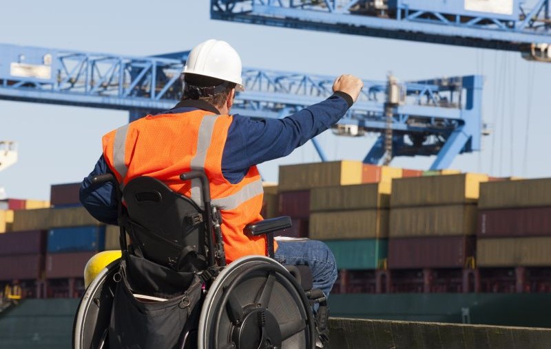 A worker using a wheelchair wearing a hard hat and orange safety vest gestures toward shipping containers and cranes at an outdoor industrial facility.