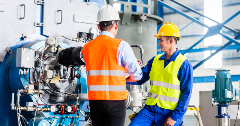 Two workers wearing safety vests and hard hats look over a piece of machinery together. One holds a clipboard, supervising the other.