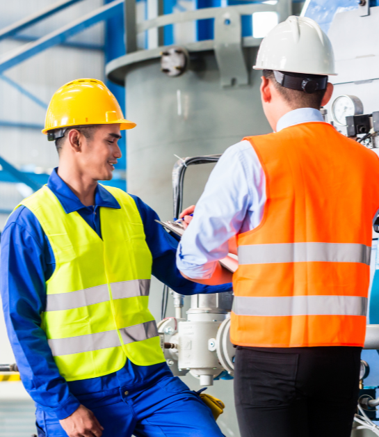 Two workers wearing safety vests and hard hats look over a piece of machinery together. One holds a clipboard, supervising the other.