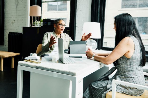 Two people sit across from each other at a desk in coversation. They both have laptops open in front of them.