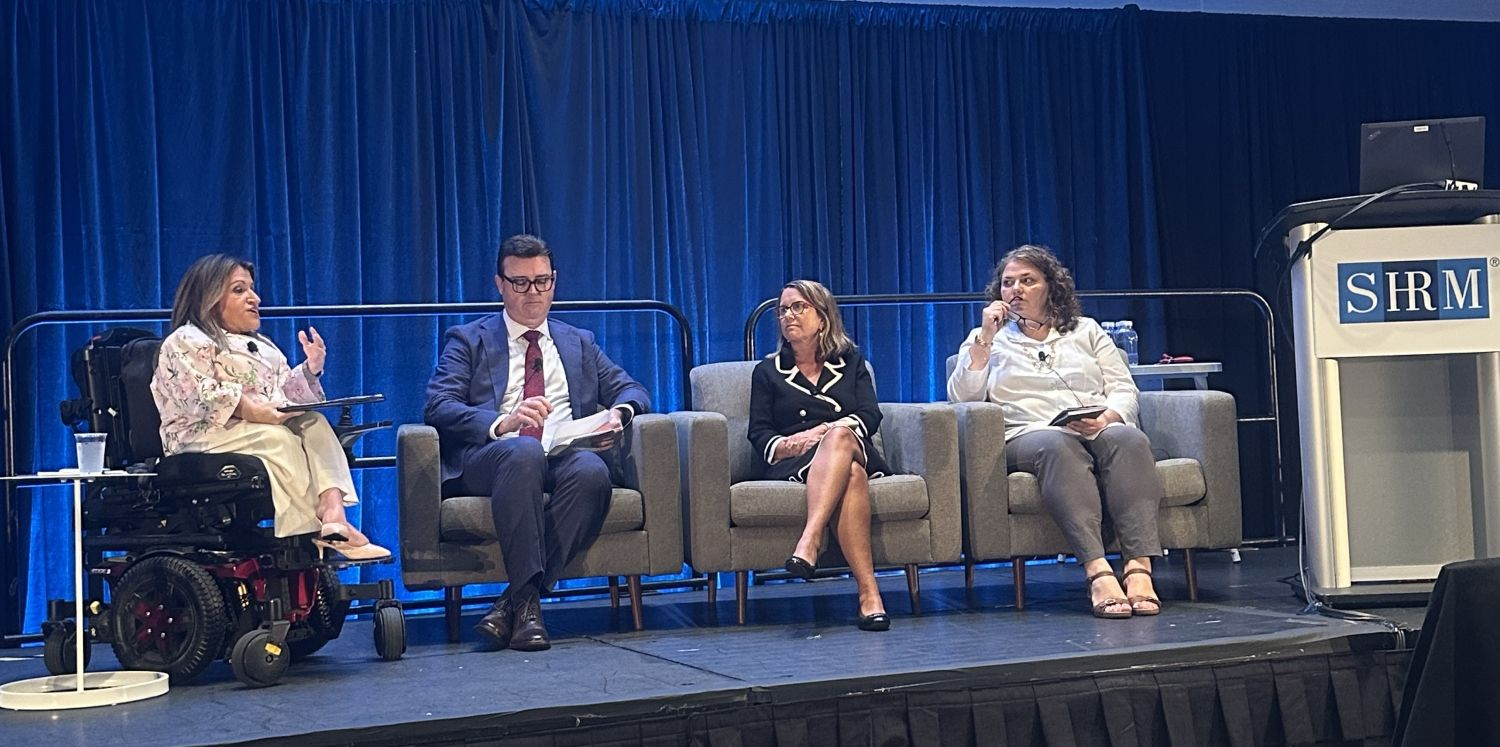 Four people sit on a stage with a blue curtain backdrop and a podium bearing the SHRM logo. The person on the far left uses a wheelchair.