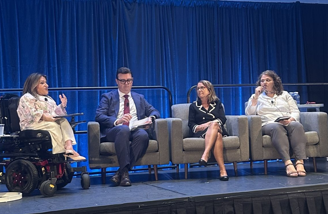 Four people sit on a stage with a blue curtain backdrop. The person on the far left uses a wheelchair.