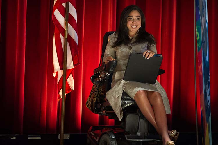 A woman in a wheelchair speaks from a stage next to an American flag