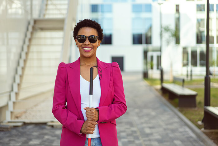 A business person who is blind smiles and stands in a courtyard while holding a cane and wearing sunglasses.