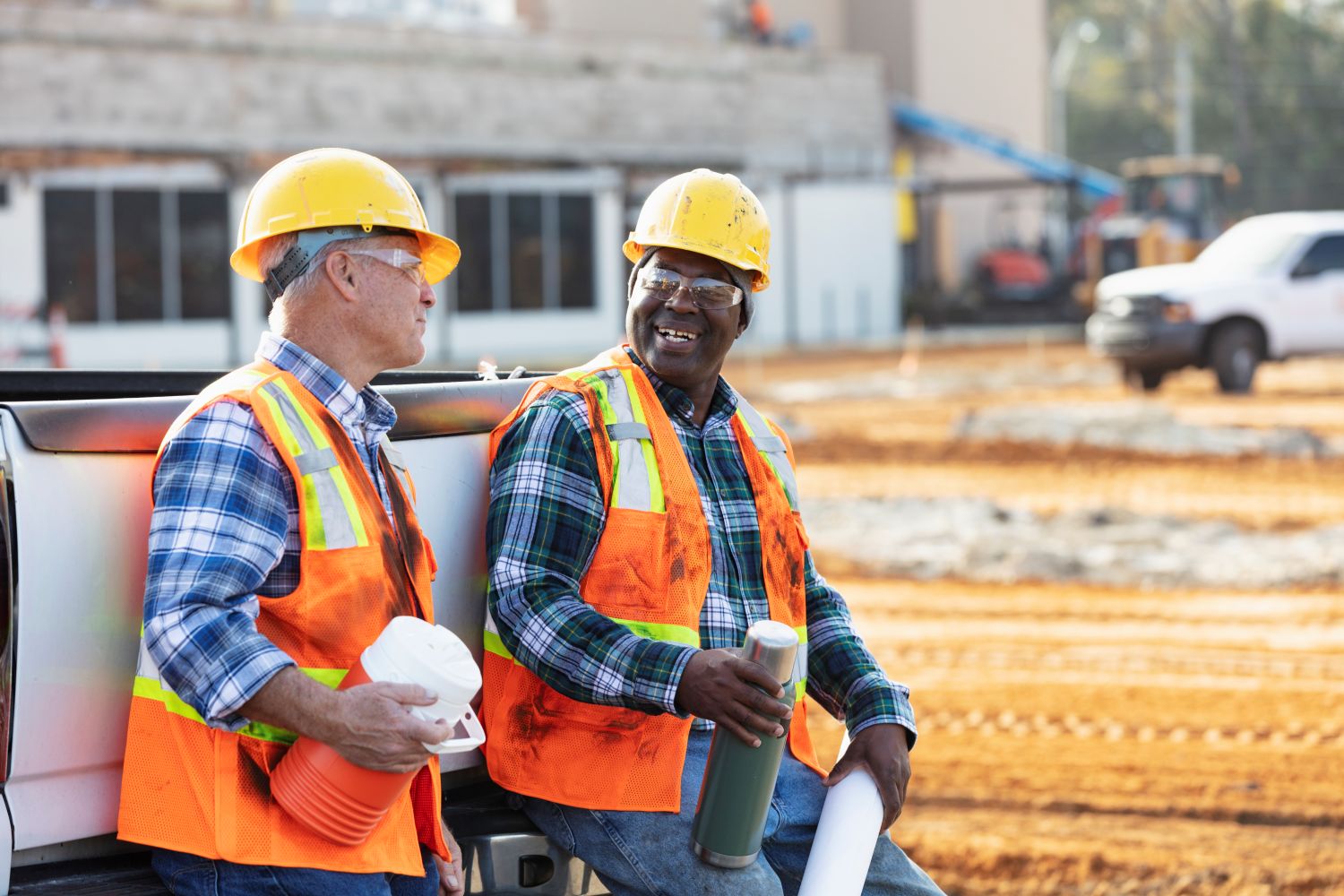 Two construction workers on a construction site taking a coffee break together. Both are wearing safety glasses, hardhats, and vests, and are leaning against a pick-up truck..