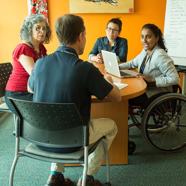 A woman in a wheelchair addresses three colleagues around a small table