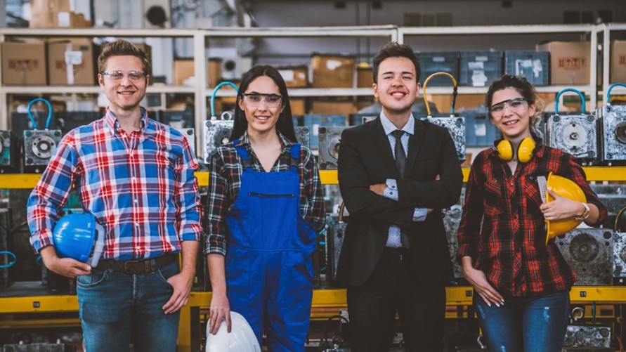 A group of four workers standing in a manufacturing workspace. They are smiling and holding safety gear.
