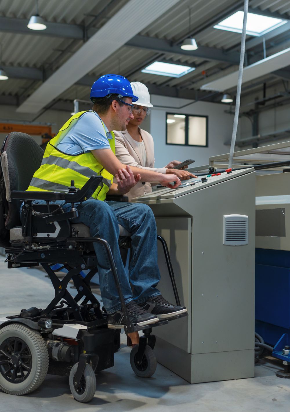A manager trains a worker how to use a machine in manufacturing facility. Both wear hard hats and the worker is sitting in an elevated electric wheelchair