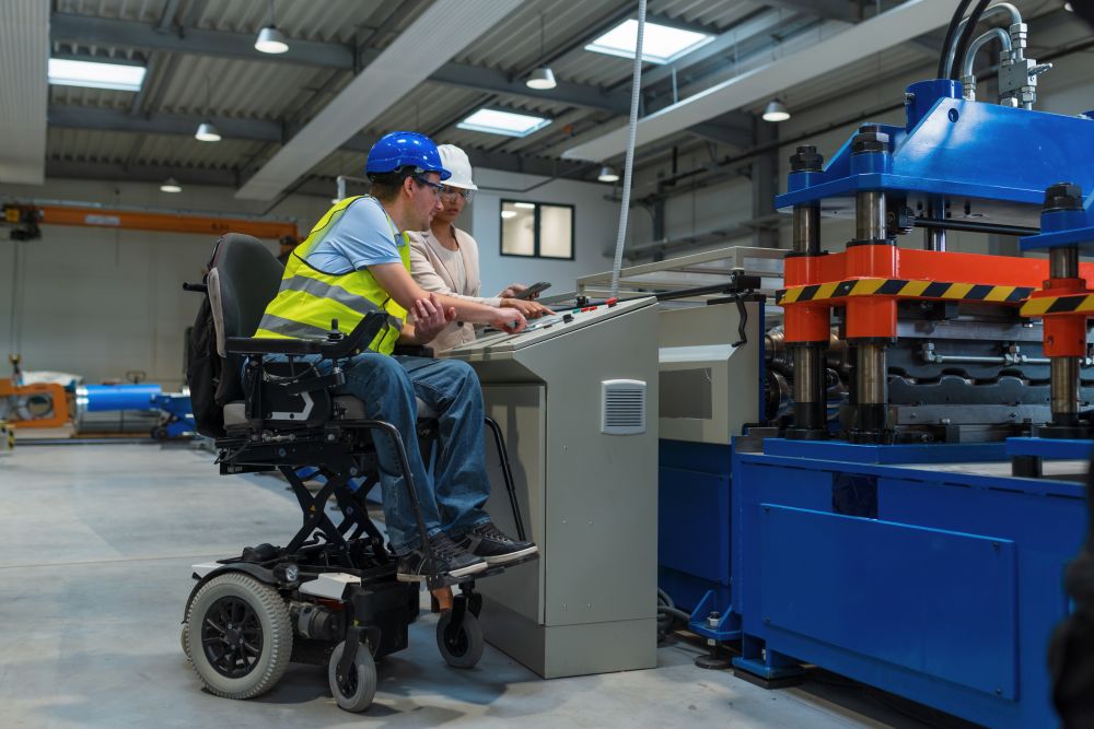 A manager trains a worker how to use a machine in manufacturing facility. Both wear hard hats and the worker is sitting in an elevated electric wheelchair