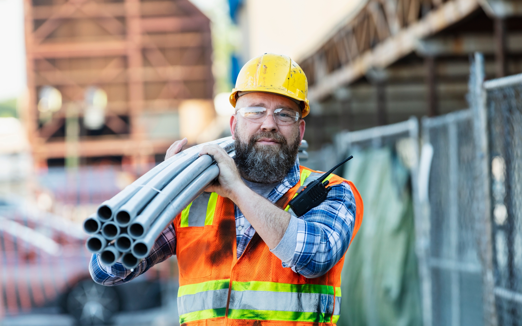 A construction worker carrying pvc pipes on his shoulder on an outdoor construction site. He is wearing a hardhat, safety glasses, and reflective vest.