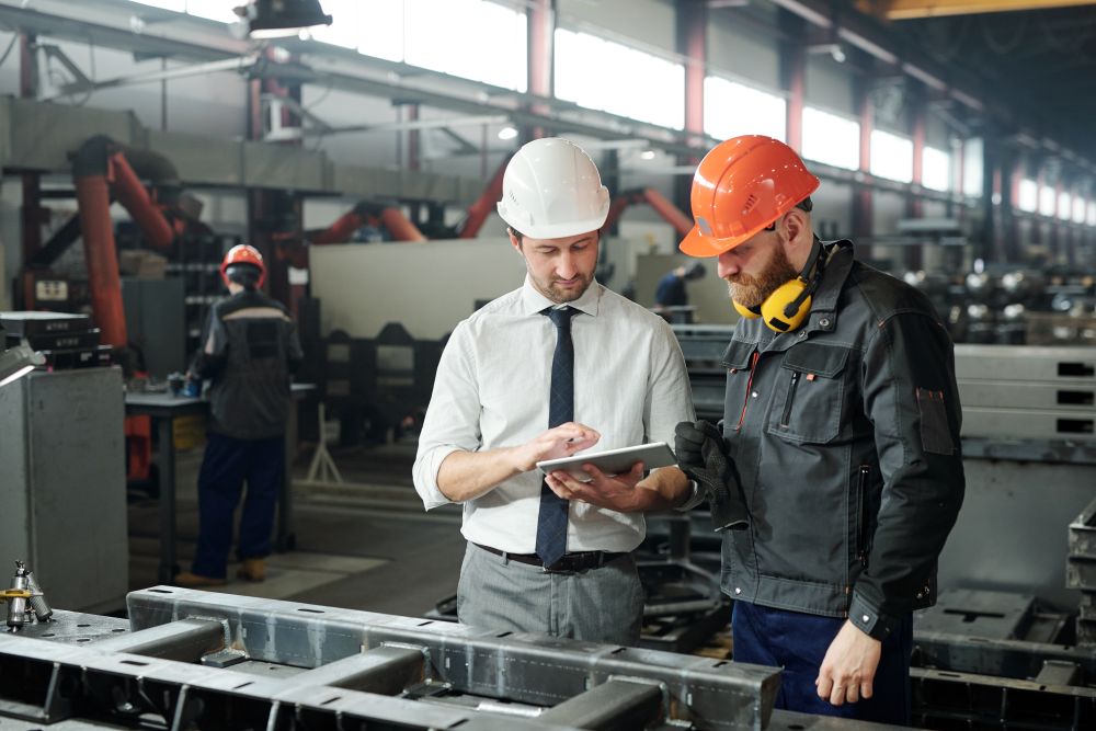 A worker in a mechanical workshop looks at his managers tablet while discussing work. Both are wearing safety equipment