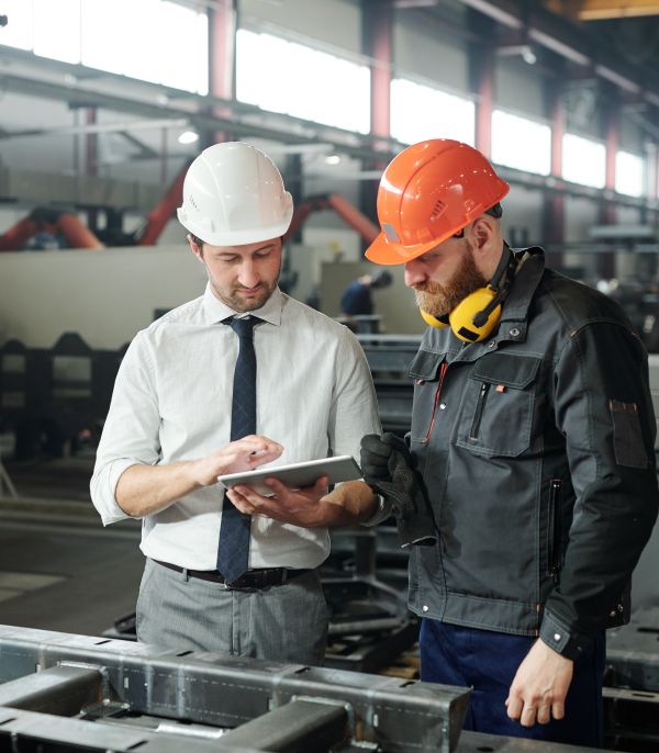 A worker in a mechanical workshop looks at his managers tablet while discussing work. Both are wearing safety equipment