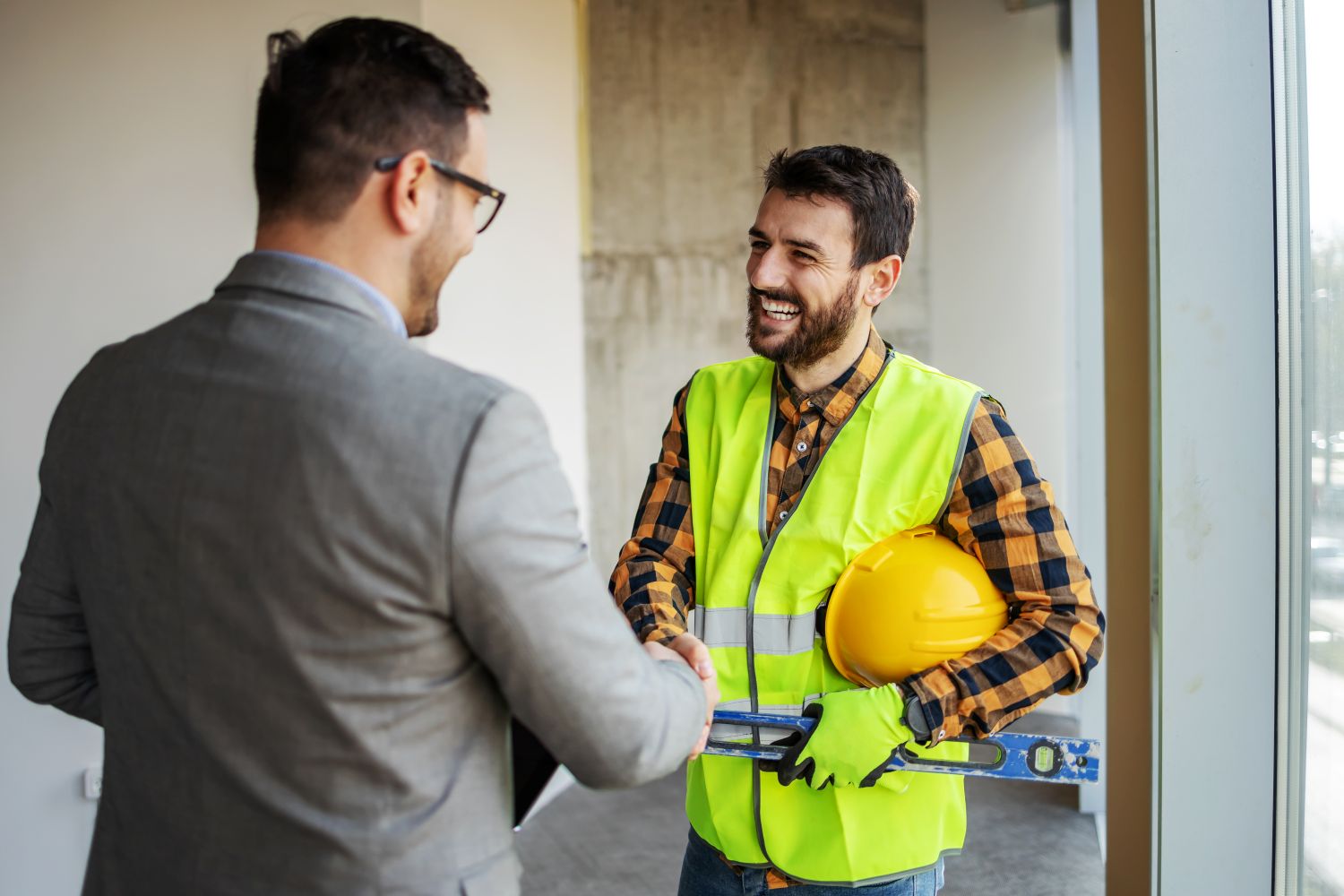 A construction worker smiling and shaking hands with his supervisor. He is wearing a safety vest and holding a hard hat and level in his other hand.