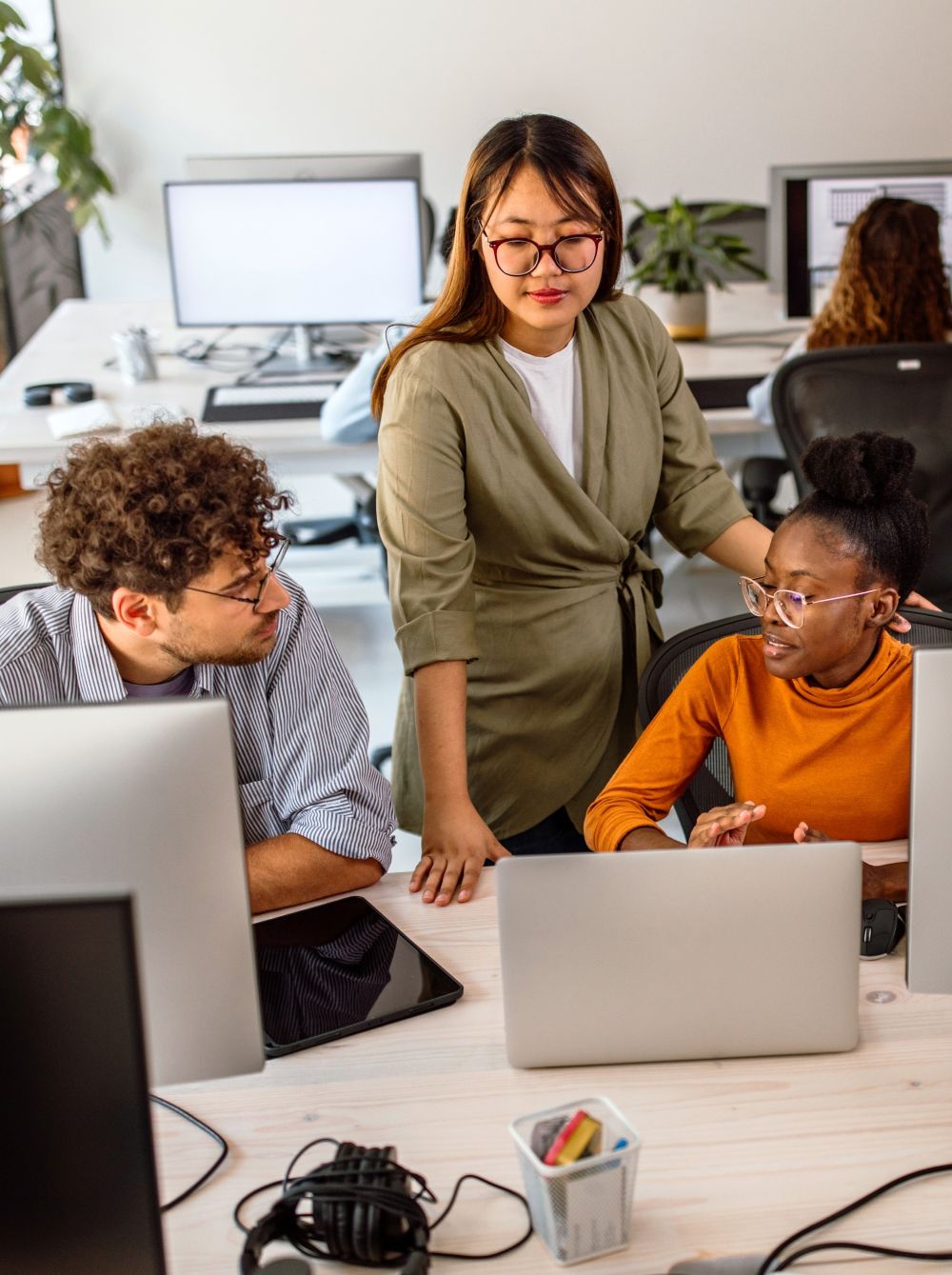 Two young tech professionals in an open concept office space sit at their computers having a conversation with another worker who is mentoring them.
