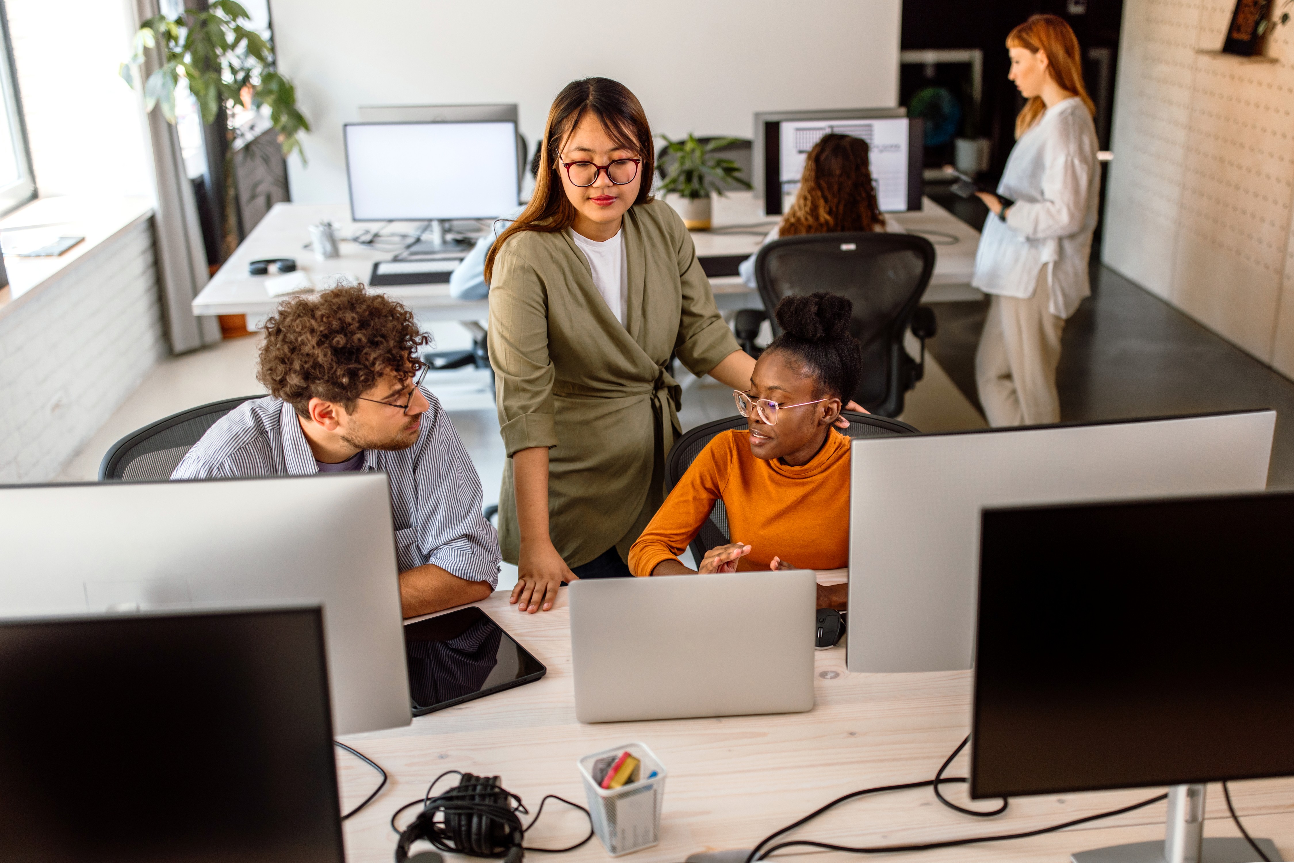 Two young tech professionals in an open concept office space sit at their computers having a conversation with another worker who is mentoring them. There is someone being assisted at their own station in the background.