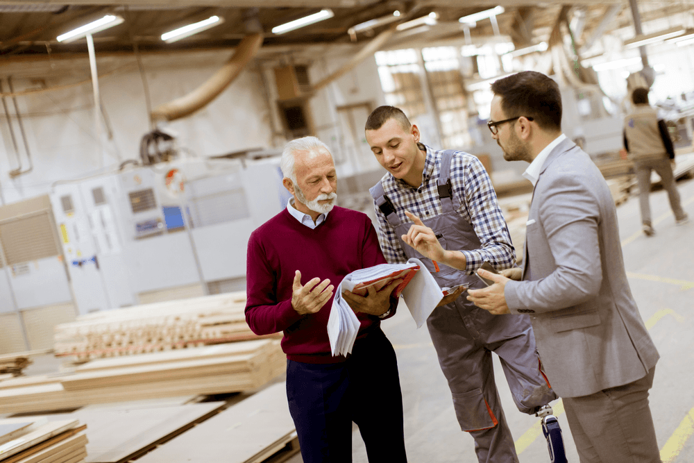 Three men working in a warehouse. An older man discusses something on a clipboard with a younger man with a crew cut and prosthetic leg. A third man holding a tablet is listening to them.
